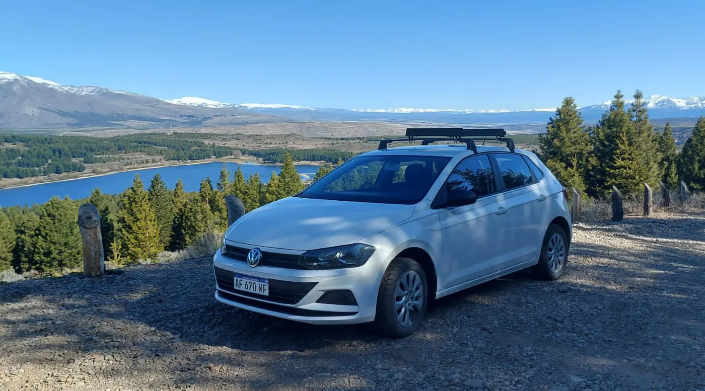 Volkswagen Polo blanco con barras portaequipaje frente a un lago y la cordillera