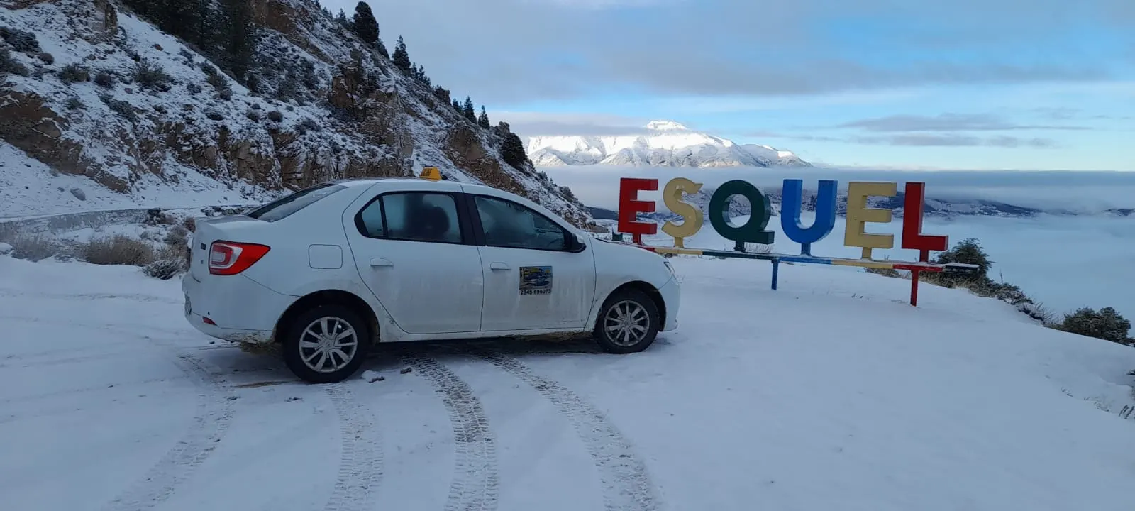 Taxi blanco junto al cartel de Esquel con nieve y la cordillera de fondo